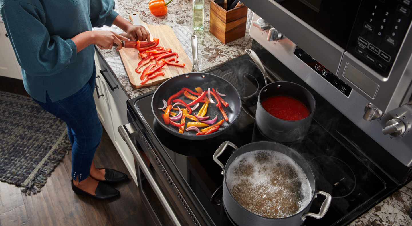 A person cutting red peppers and adding them to a frying pan next to a pot of simmering water on a Maytag® electric stovetop