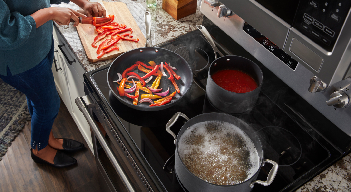 Overhead shot of woman cutting peppers with peppers, sauce & noodles boiling on the cooktop next to her