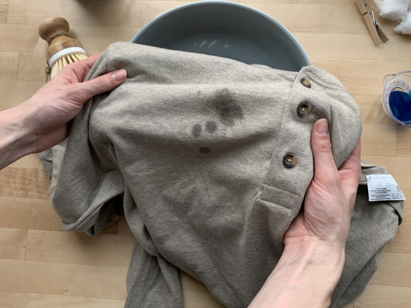 Gray shirt with grease stains and a cup of blue detergent next to it