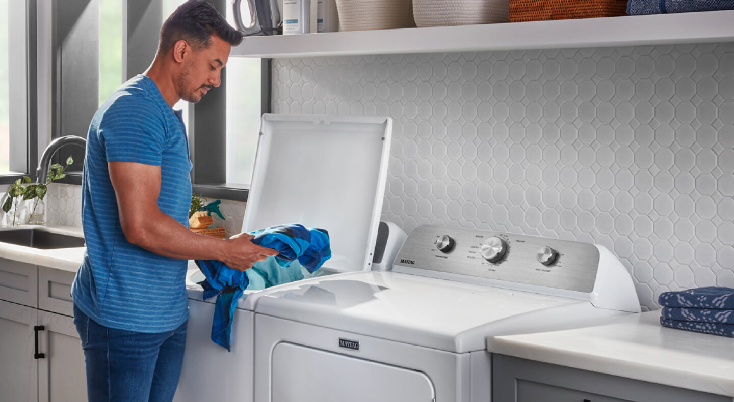 A person adding laundry items to a Maytag® top load washer in a modern laundry room
