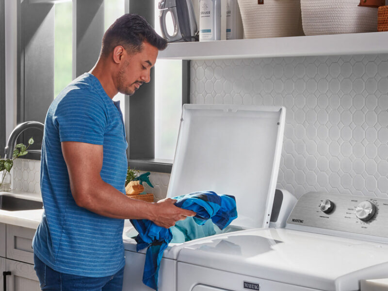 A person adding laundry items to a Maytag® top load washer in a modern laundry room
