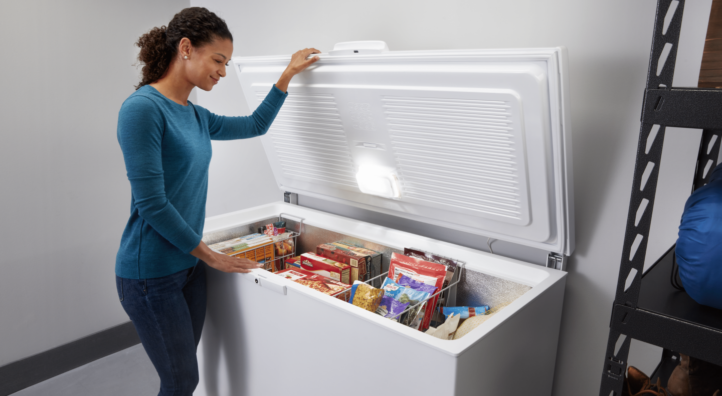 A woman opening a white chest freezer with various frozen items inside