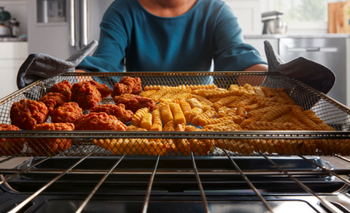 Person pulling an air fryer basket with cooked chicken nuggets and crinkle cut fries out of an oven