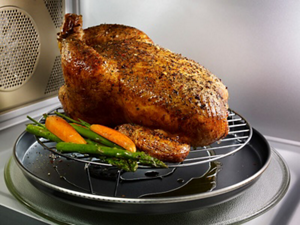 A whole chicken with vegetables cooking on a grilling accessory inside of a microwave