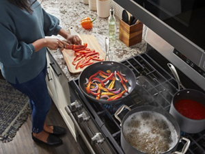 An overhead view of a person chopping vegetables next to a Maytag® gas cooktop with a frying pan and boiling pots
